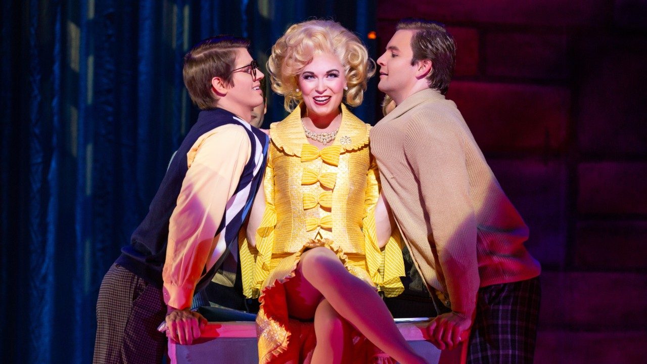  A blonde white woman with 60's hair and yellow dress grabs two white men by the collars and holds them close to her. She's sitting crosslegged on a desk. The two men look uncomfortable but excited.