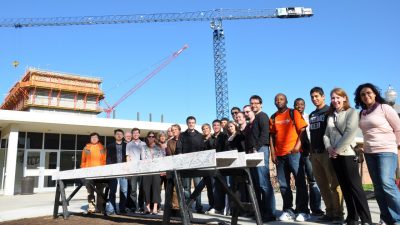 Virginia Tech faculty, staff, and students gather to sign one of the beams used in construction at the Moss Arts Center. Behind them is a clear blue sky and two cranes.