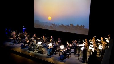 The Philip Glass Ensemble, seen on stage from an upper level box seat, performs at the Moss Arts Center during the inaugural performance. A large projection screen shows a setting sun over an African savanna.