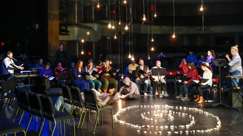  Image during (Be)longing performance at the Moss Arts Center. Performers sit on the Fife stage in a semicircle with music stands in front of them. There are hanging lights coming down from above the stage, there are rocks in a spiral shape on the stage floor.