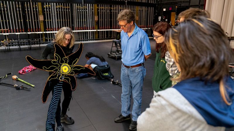experiences_lightwire-theater_class-visit_lev-birman Virginia Tech students in a School of Performing Arts lighting design class meet with members of Lightwire Theater to learn tricks of the trade. A white woman with shoulder length blonde hair holds a light-up flower puppet and explains her creative process to three students and a professor.