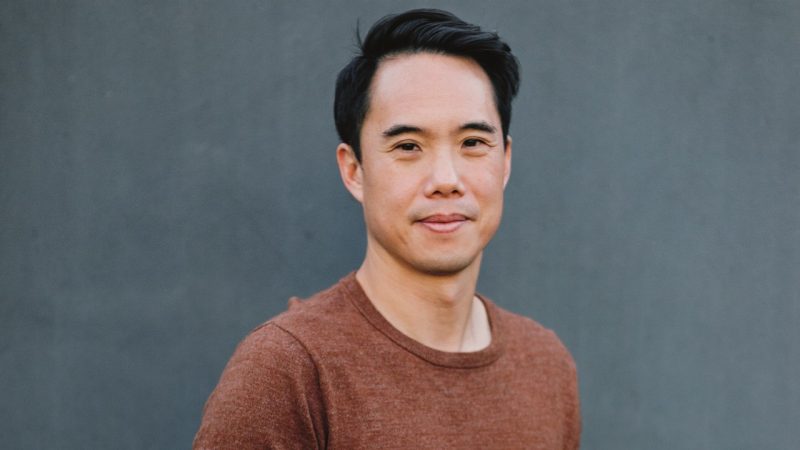 Author Charles Yu, a young Asian man with short dark hair, wears a rust brown T-shirt and smiles towards the camera in front of a medium grey background.