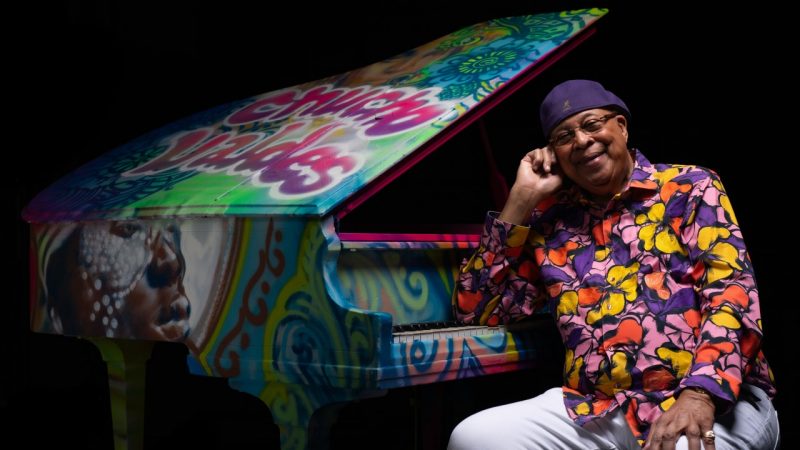 Cuban jazz pianist Chucho Valdes, an older Hispanic man, wears a purple hat and a multicolored floral button down shirt. He sits at an open grand piano covered from top to bottom in spray paint art and smiles towards the camera.
