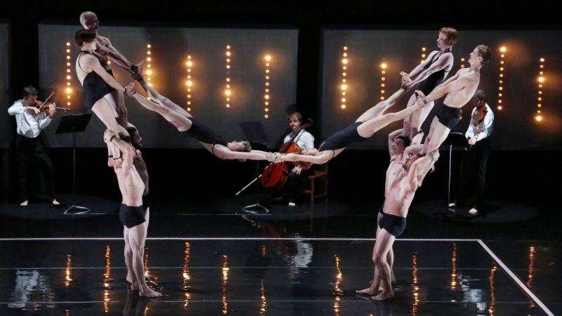 The cast of Circa's "Opus" perform on stage wearing black leotards or pants. Four people stand on the shoulders of four others, while the pairs each hold a person's feet. The two people, upside down, clasp hands in the center. A string quartet plays in the background.