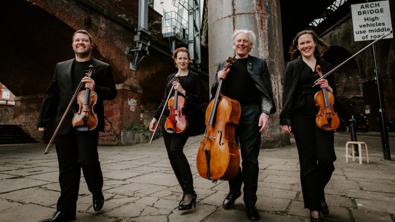 Members of the Dante Quartet hold their instruments in an industrial area of a city and smile widely.