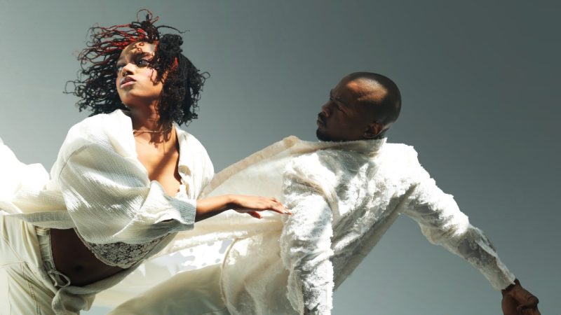 Jamaal Bowman, right, and Keturah Stephen, left, dancers with A.I.M by Kyle Abraham, wear all white and appear to gall together across the frame in front of a light grey background.