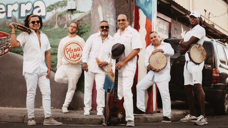The members of Plena Libre, six Hispanic men, wear all white and hold their instruments on a city street.