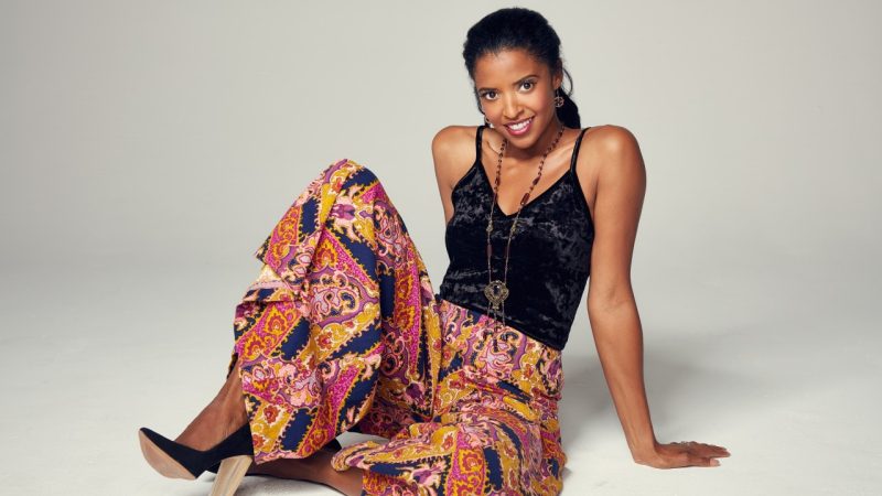 Broadway star Renée Elise Goldsberry, a young Black woman, sits in pink and purple paisley widelegged pants and a black velvet tank top and smiles towards the camera.