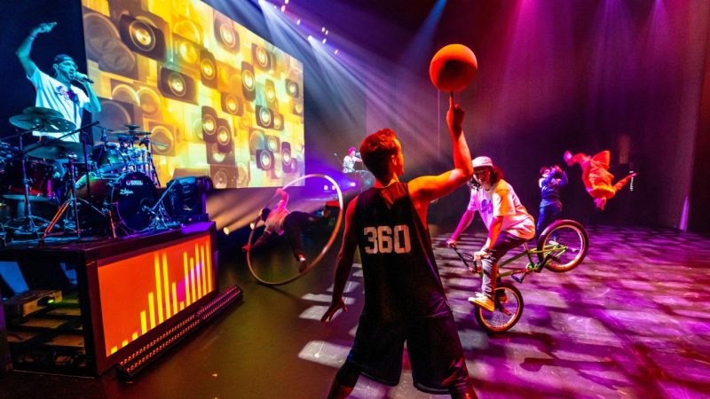 Members of 360 ALLSTARS perform on stage, on DJing, one balancing a basketball on a finger, one balancing on one wheel of a BMX bike.