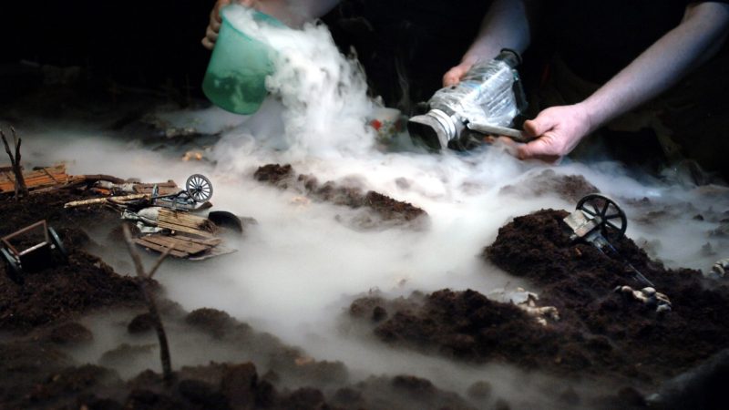 A member of Hotel Modern uses a video recorder to capture a minature set of a World War I battlefield draped in fog, made from dry ice.