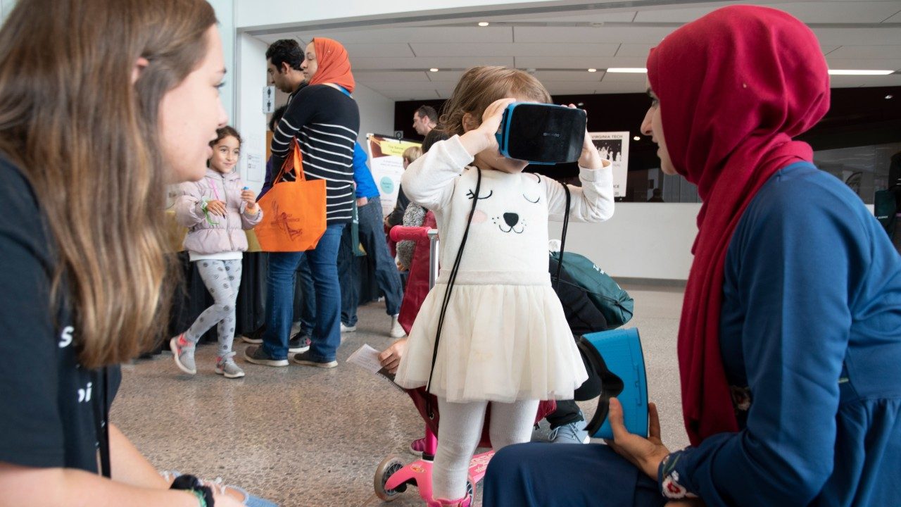  Young girl in white skirt and shirt with an animal's face holds a viewfinder up to her face. Two women are kneeling in front of her. This is in the Moss Arts Center's grand lobby during Science Festival.