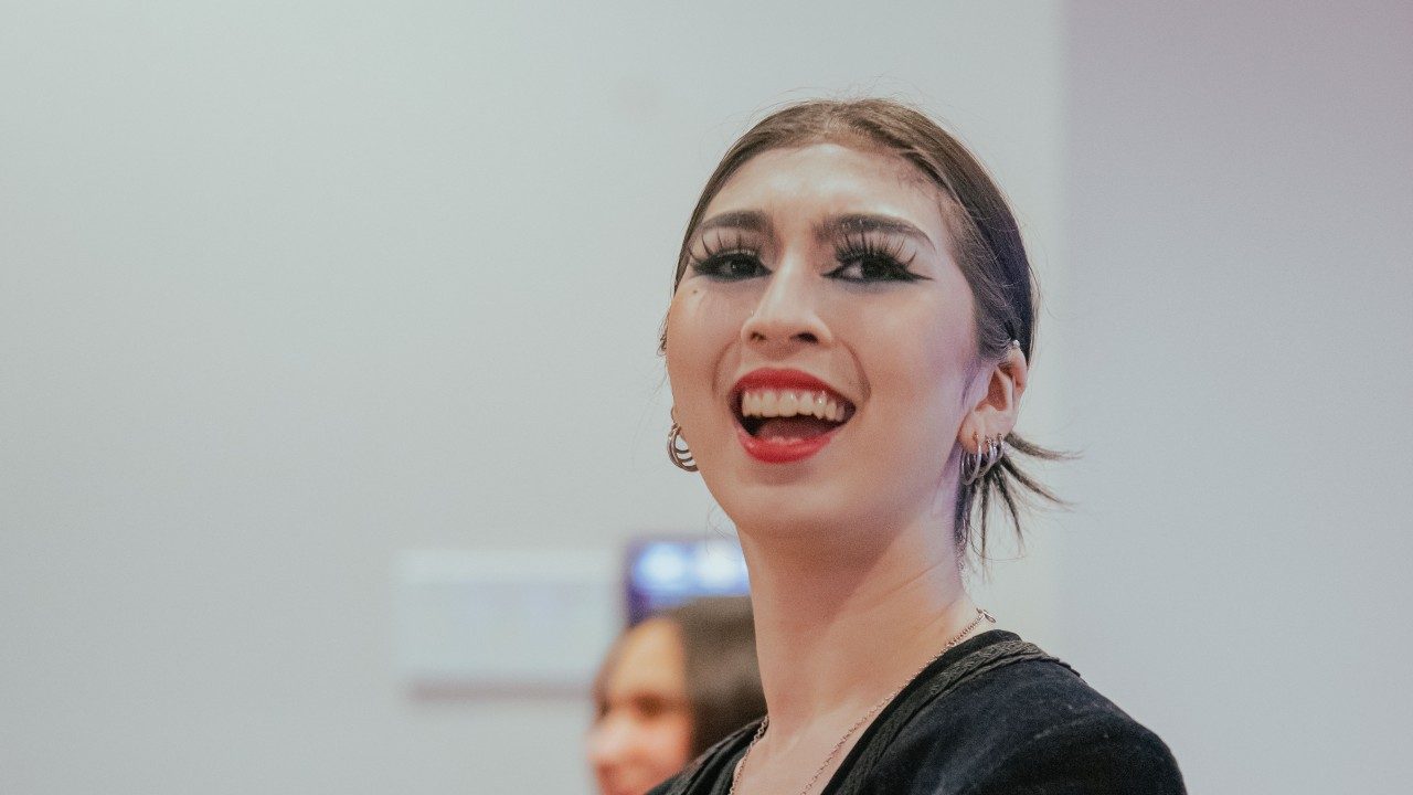  A young woman with dark brown hair pulled back into a bun smiles widely. She wears a bold red lipstick and graphic black eyeliner.