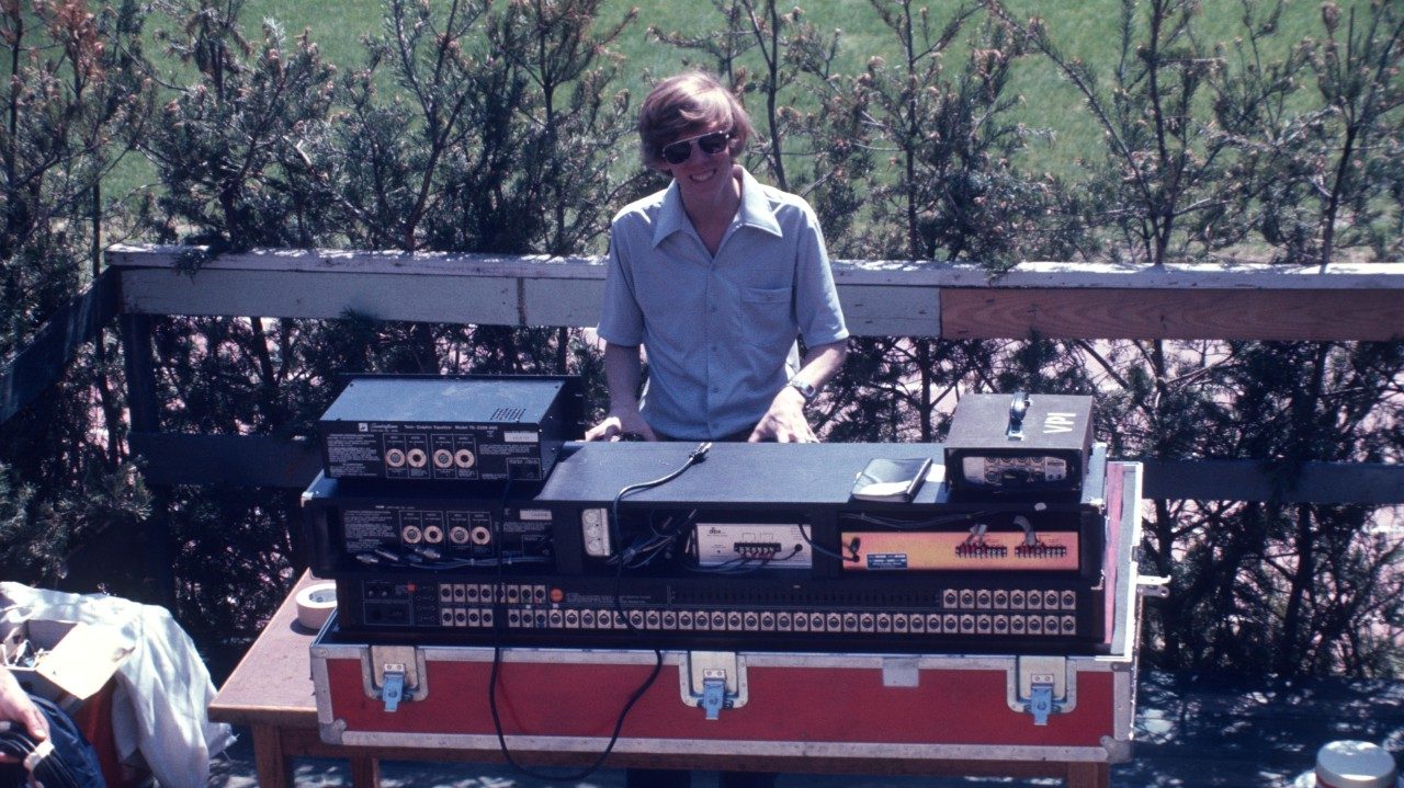  A member of the New Virginians, a white man with shaggy light brown hair and sunglasses, stands at a mixing board outside, a line a evergreen trees directly behind him. 