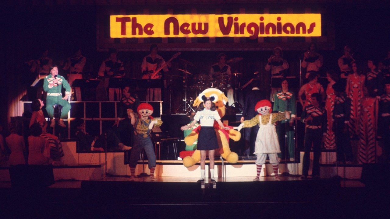  Members of the New Virginians perform onstage. Behind the group is a large, light-up sign with a yellowy-orange background and maroon letters spelling "The New Virginians." Some members in costumes (Raggedy Anne and Raggedy Andy, Winnie the Pooh, and a woman in a black shirt, white shirt, and black mouse ears with pigtails). Nearby, a man seated on a stool and holding a microphone is under a spotlight. He wears a green shirt and pants. 