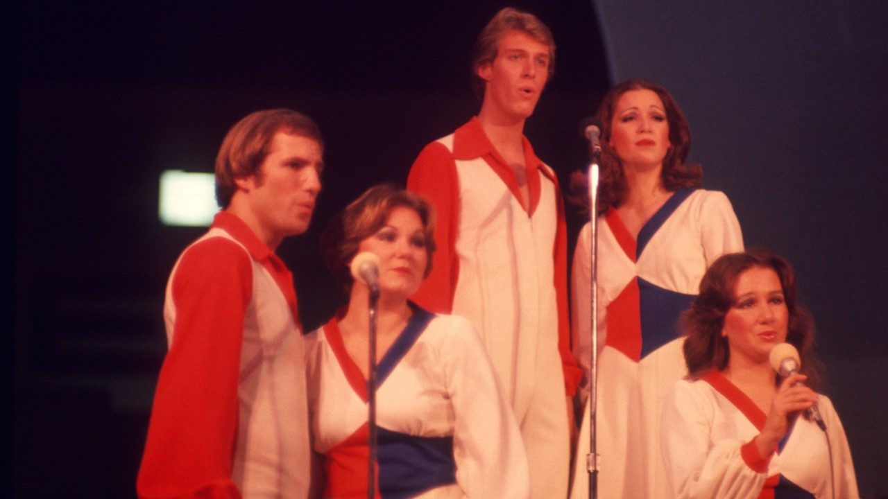  Five members of the New Virginians perform on stage. From left, a white man with medium length light brown hair, a white woman with short strawberry blonde hair, a white man with short blonde hair, a white woman with long curled reddish brown hair, and a white woman with long dark brown hair.  The men wear white jumpsuits over red button down shirts, and the women wear white dresses with red and blue accents at the waist and neckline.