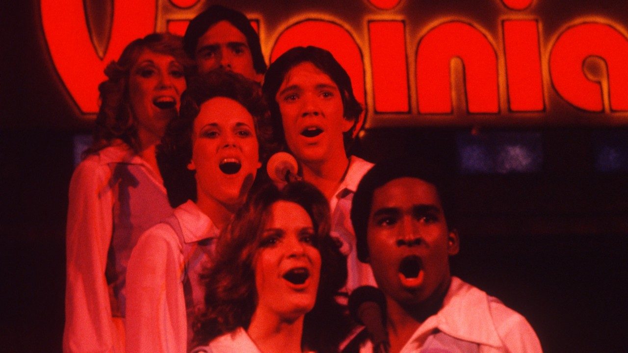  Three pairs of New Virginians sing into shared microphones under red lighting, a red, light-up "Virginia" visible behind them. From the front is a white woman and a Black man, a white woman and a white man, and a white woman and a white man.