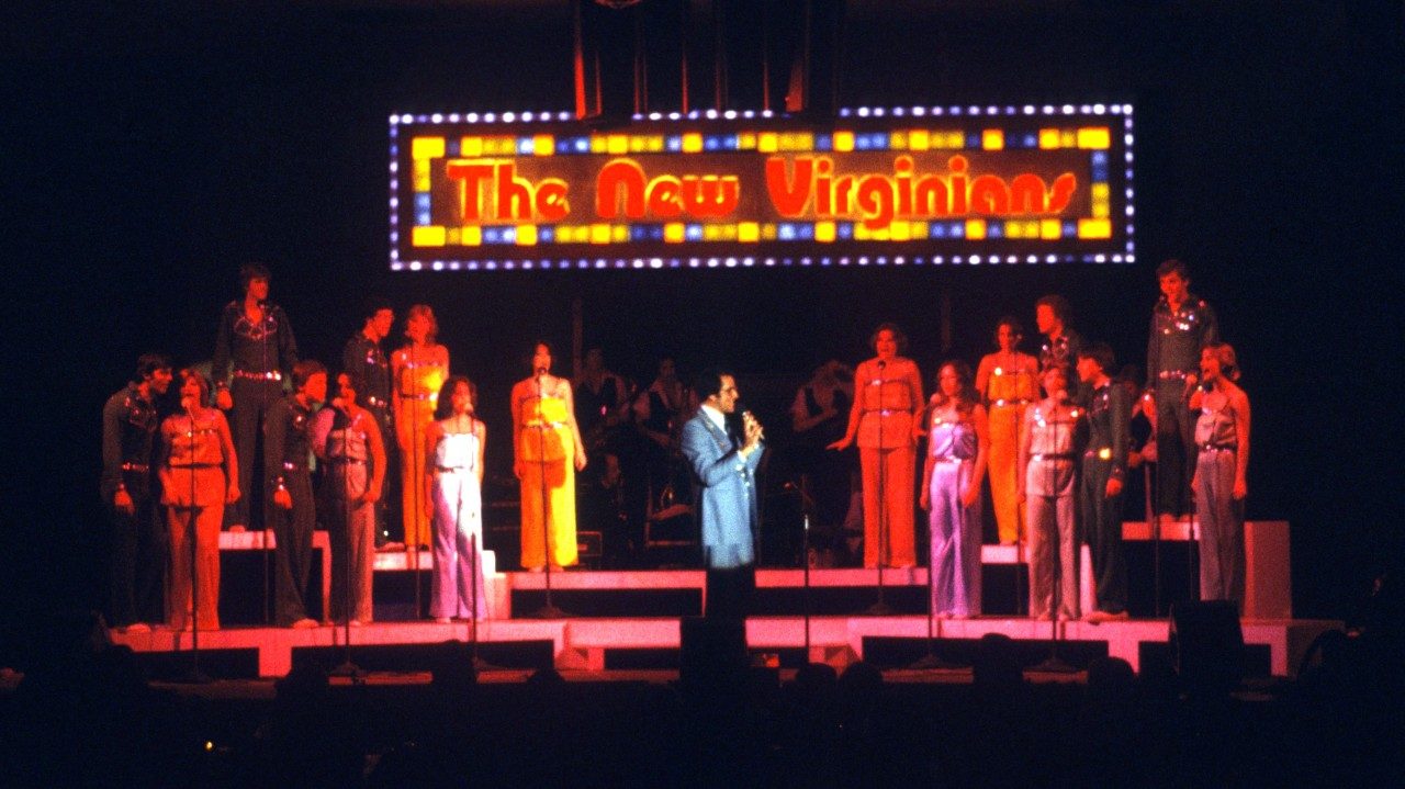  The New Virginians perform on stage in multicolored costumes. In the middle is a white man with dark brown hair and a light blue suit. Behind them all is a light up sign that reads "The New Virginians"